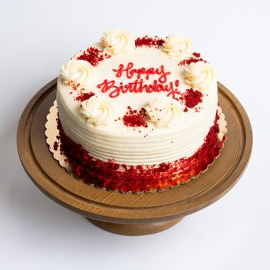 Red Velvet Birthday Cake from Mia's Bakery featuring a round single-layer cake with white cream cheese frosting and horizontal ridged texture on the sides. The birthday cake has red velvet cake crumbs pressed around the bottom edge and is decorated with piped vanilla buttercream rosettes around the top edge, each sprinkled with red velvet cake crumbs. 'Happy Birthday!' is written in red cursive frosting in the center. Presented on a wooden pedestal cake stand against a white background.