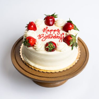 Strawberry Shortcake Birthday Cake from Mia's Bakery featuring a round single-layer cake covered in smooth white vanilla buttercream with horizontal ridged texture on the sides. The birthday cake is decorated with alternating fresh strawberries and piped white buttercream rosettes around the top edge, with 'Happy Birthday!' written in red cursive frosting in the center. Presented on a wooden pedestal cake stand against a white background.