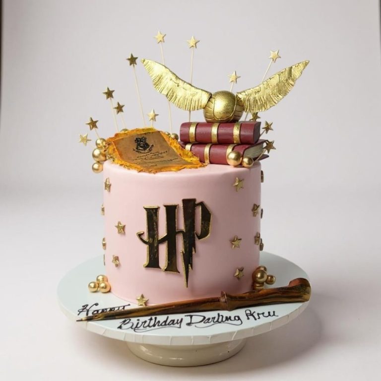 Pink Harry Potter–themed cake decorated with gold HP logo, edible golden snitch with wings, stacked red books, golden stars, wand, and parchment scroll accents