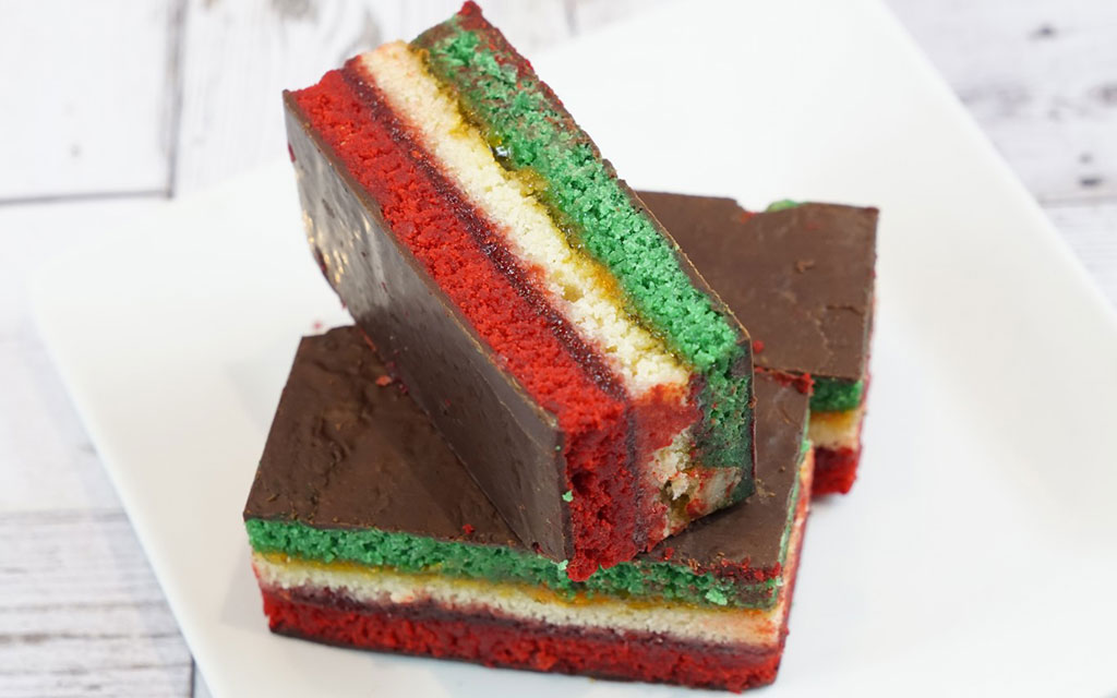 Italian rainbow cookies with red green and white layers topped with chocolate ganache on white plate christmas dessert