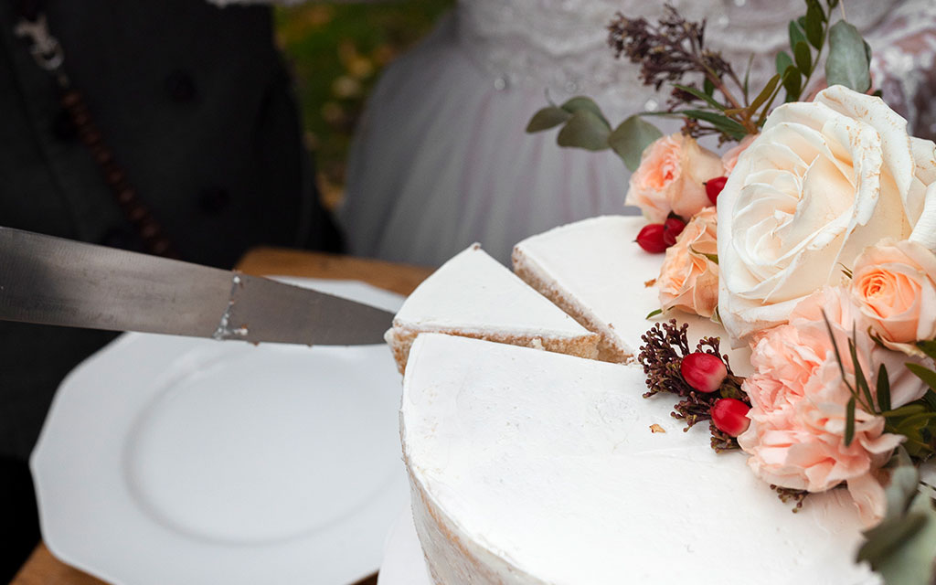 Couple cutting white spring wedding cake decorated with fresh peach roses and red berries at reception celebration