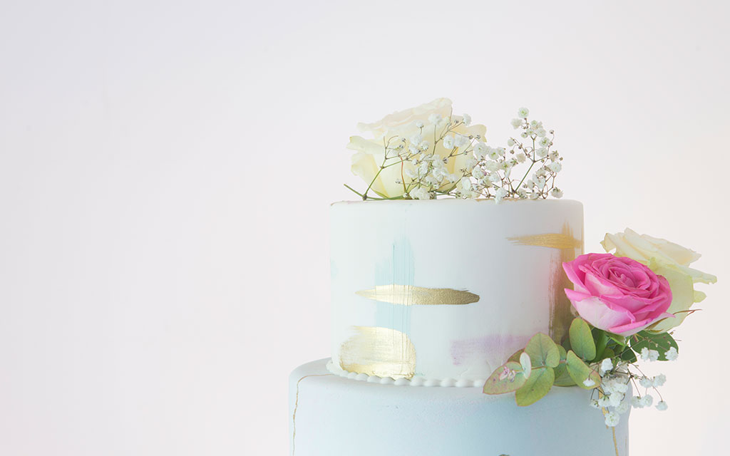 Two-tier spring wedding cake with hand-painted watercolor design, gold brushstrokes, fresh pink rose, and baby's breath