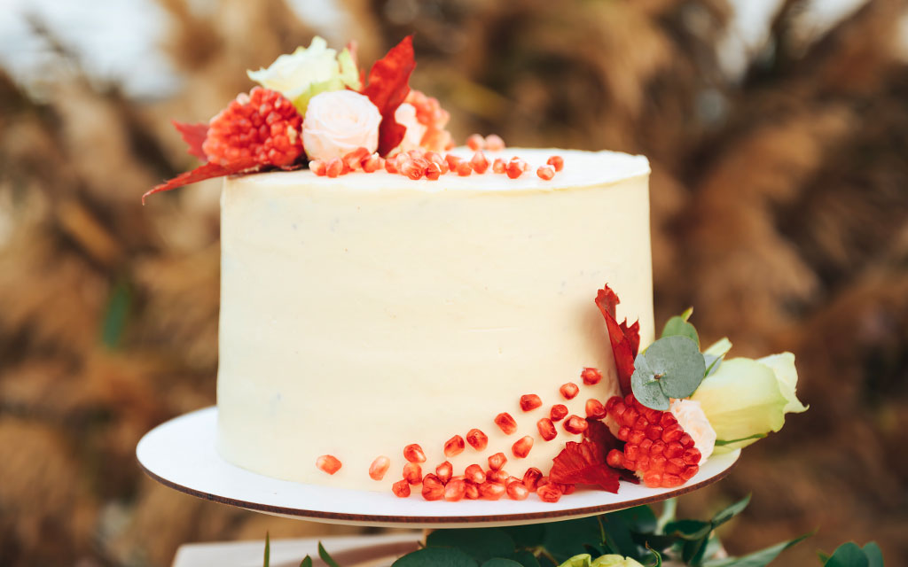 Fall wedding cake with smooth buttercream, pomegranate seeds, and red autumn flowers on gold stand