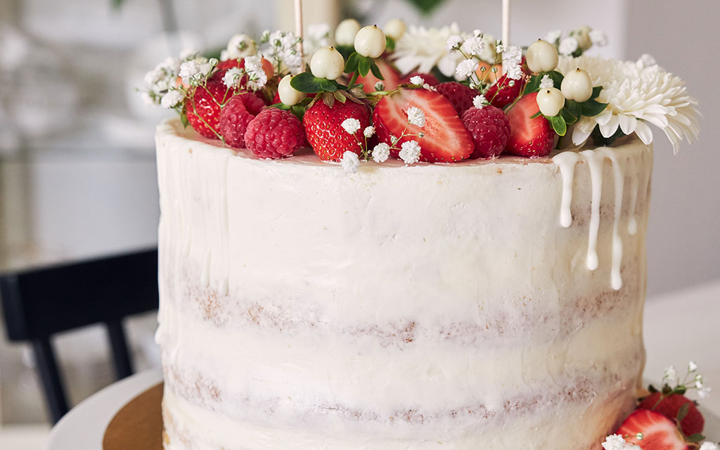 Semi-naked wedding cake with white drip, fresh strawberries, raspberries, and white flowers on top