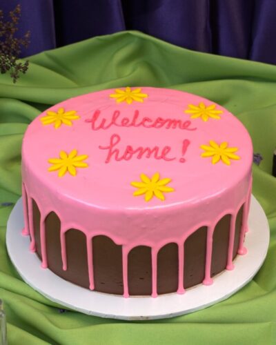 Round chocolate cake with a pink drip, yellow fondant flowers and "Welcome home!" piped on top, Mia's Bakery