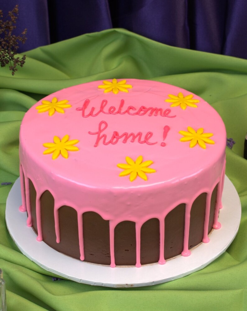 Coraline Cake, Mia's Bakery Round chocolate cake with a pink drip, yellow fondant flowers and "Welcome home!" piped on top, Mia's Bakery