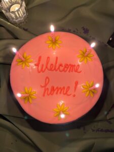 Top view of a pink cake with "Welcome home!" piped on top and yellow flowers with lit candles, Mia's Bakery