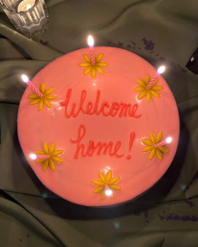 Top view of a pink cake with "Welcome home!" piped on top and yellow flowers with lit candles, Mia's Bakery