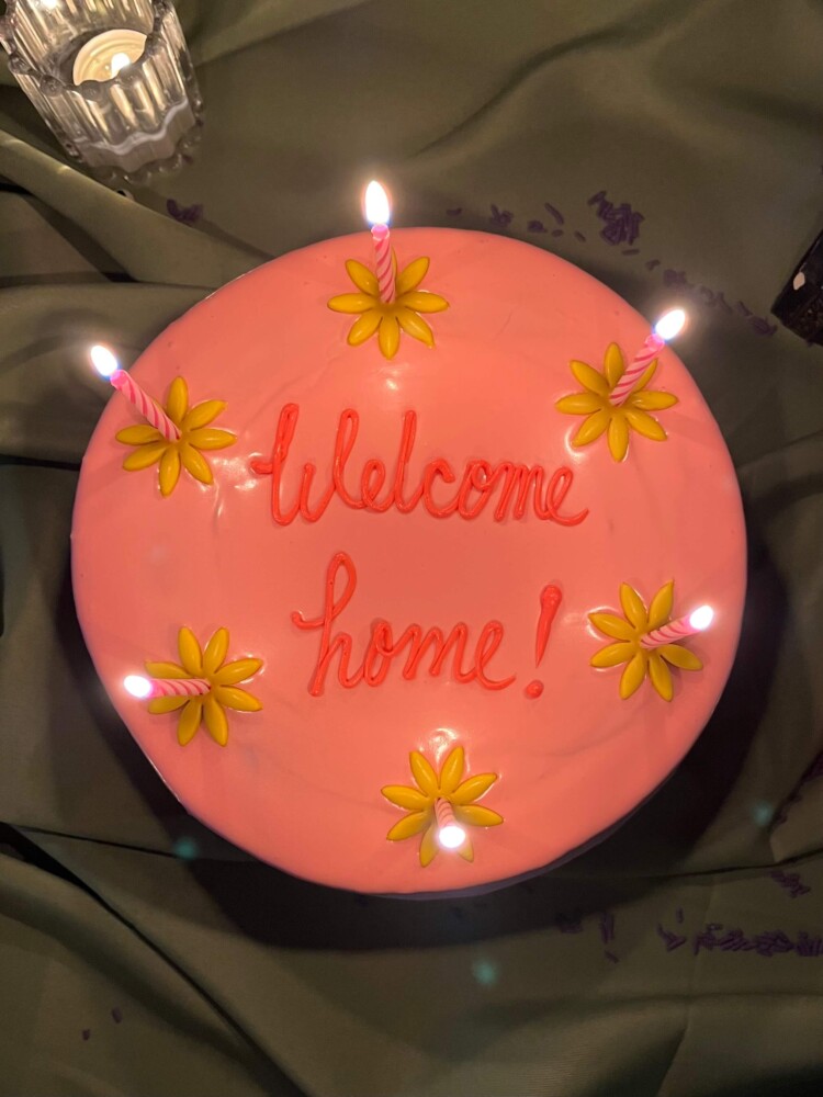 Coraline Cake Top View, Mia's Bakery Top view of a pink cake with "Welcome home!" piped on top and yellow flowers with lit candles, Mia's Bakery