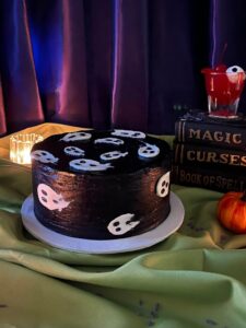 Ghosts Cake in black frosting with white ghosts, beside spell books and a small pumpkin, Mia's Bakery