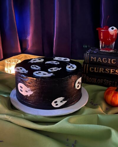 Ghosts Cake in black frosting with white ghosts, beside spell books and a small pumpkin, Mia's Bakery