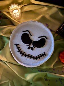 Top view of a white buttercream cake with a grinning skeleton face piped in black, Mia's Bakery