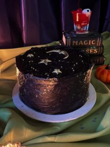 Midnight Moon Cake in black frosting with a gold moon and stars, beside spell books and a pumpkin, Mia's Bakery