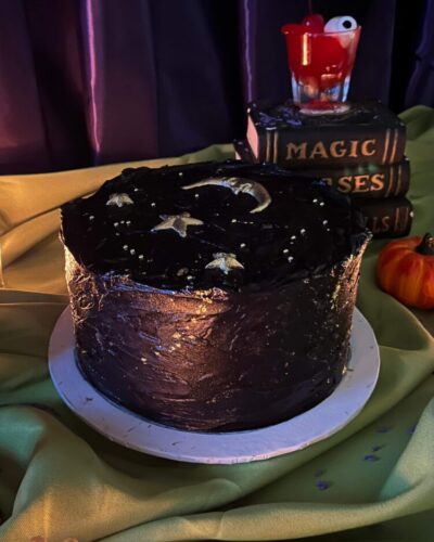 Midnight Moon Cake in black frosting with a gold moon and stars, beside spell books and a pumpkin, Mia's Bakery