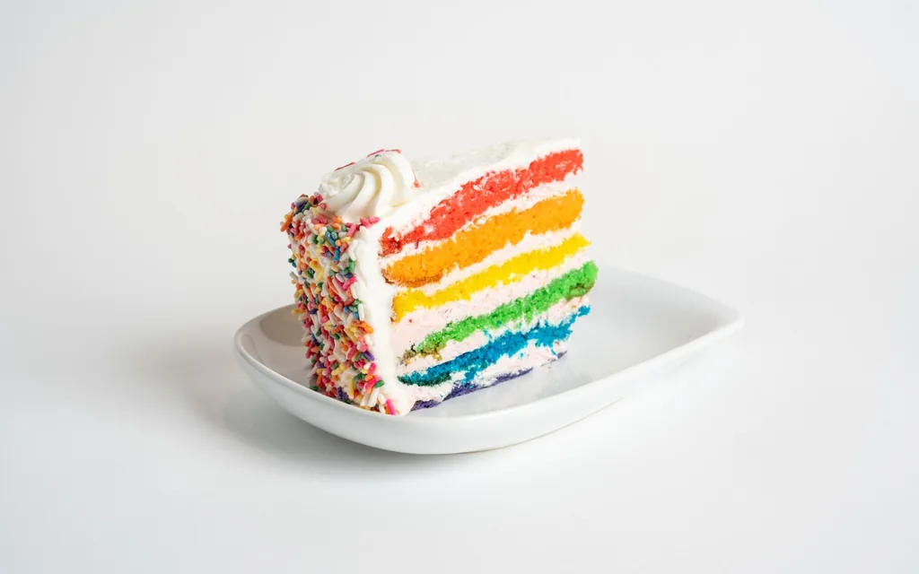 Slice of rainbow cake with six colored vanilla layers, strawberry buttercream and a coat of rainbow sprinkles